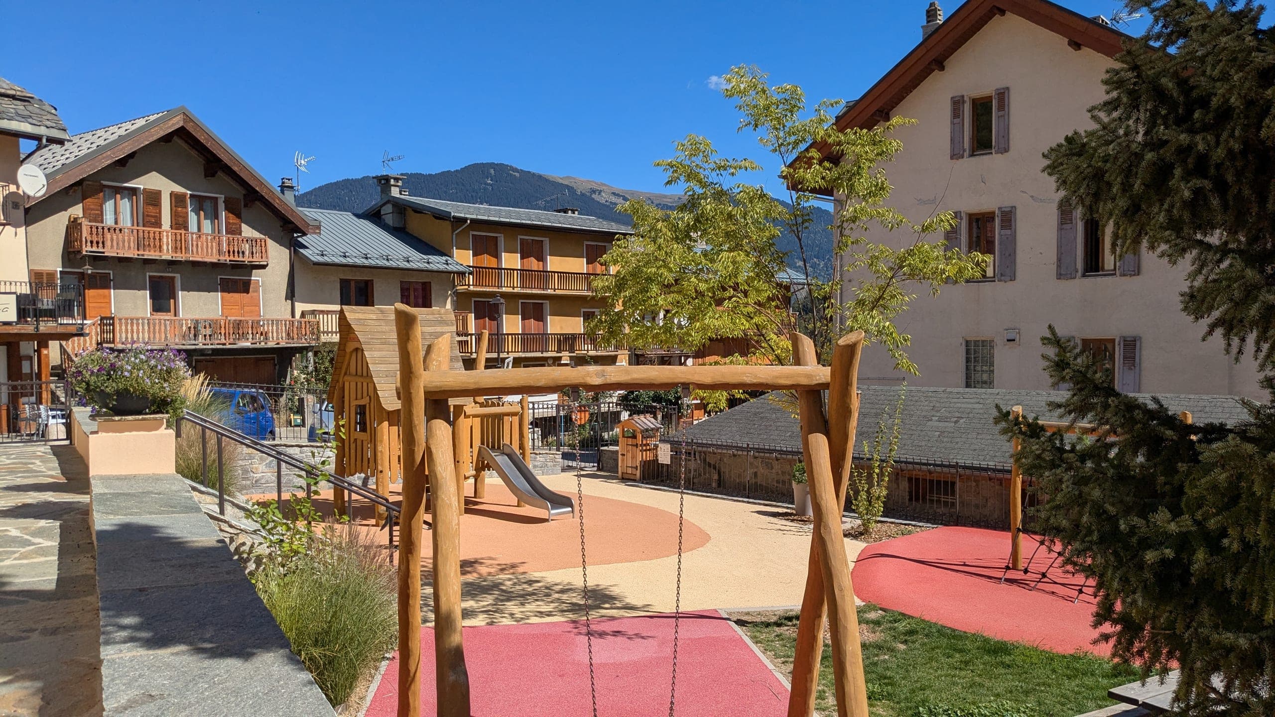 L’aire de jeux terminée en vue large : structures en rondins de bois massif sur sol souple rouge et ocre, portique de balançoire, jeune arbre planté, massifs de graminées et murets de pierre, chalets et façades enduites du village tout autour, montagne boisée au fond.