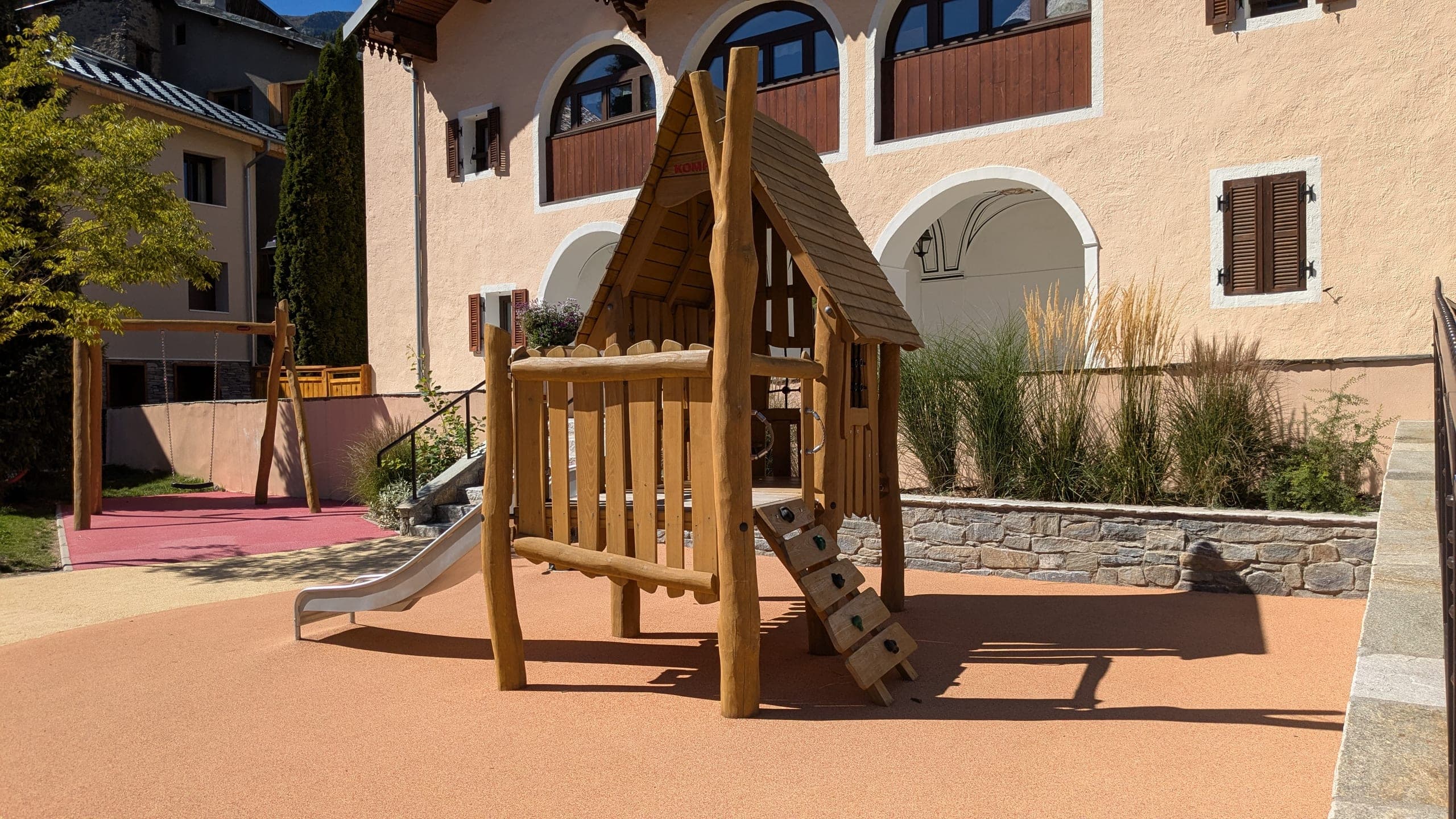 Une cabane de jeu en bois à toit pentu, avec toboggan métallique et échelle d’escalade à prises, posée sur le sol souple ocre devant une grande façade rose pâle à arcades ; graminées hautes et muret de pierre à droite, balançoire au second plan.