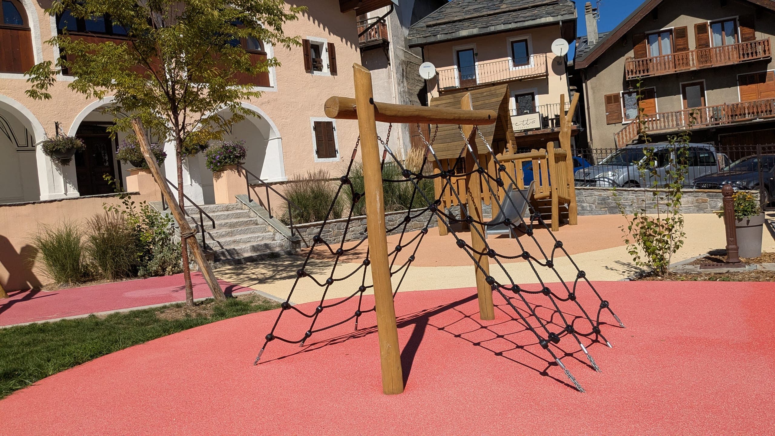 Un filet d’escalade tendu entre deux poteaux de rondins sur le sol souple rouge ; derrière, la cabane et son toboggan, un mail de jeunes arbres, les façades à arcades du village et les voitures stationnées.
