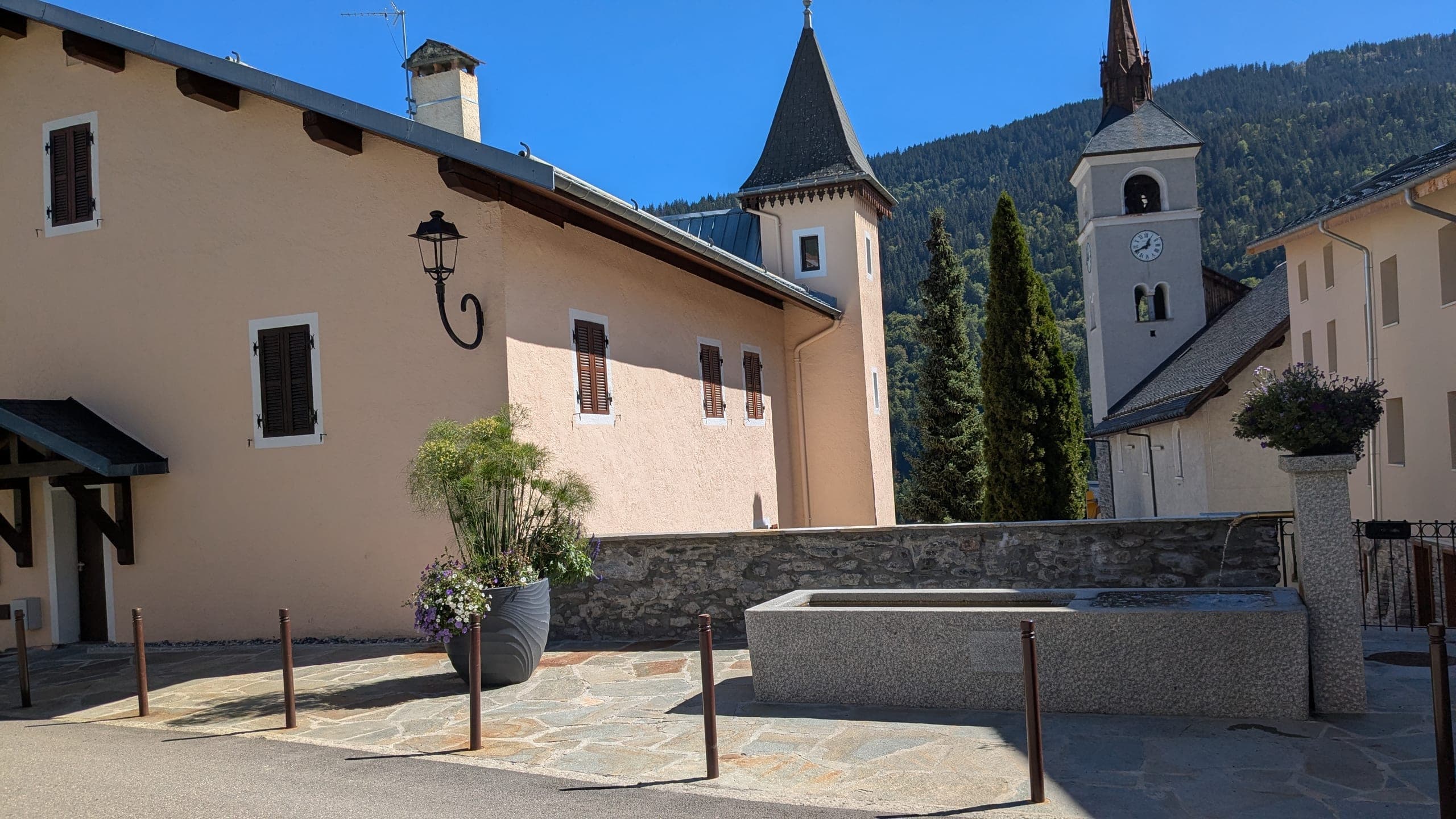 La place du village après travaux : la fontaine de granit et son eau courante, le dallage de pierre et ses potelets, le mur bas en pierre, le clocher et son horloge derrière, la montagne boisée au fond.