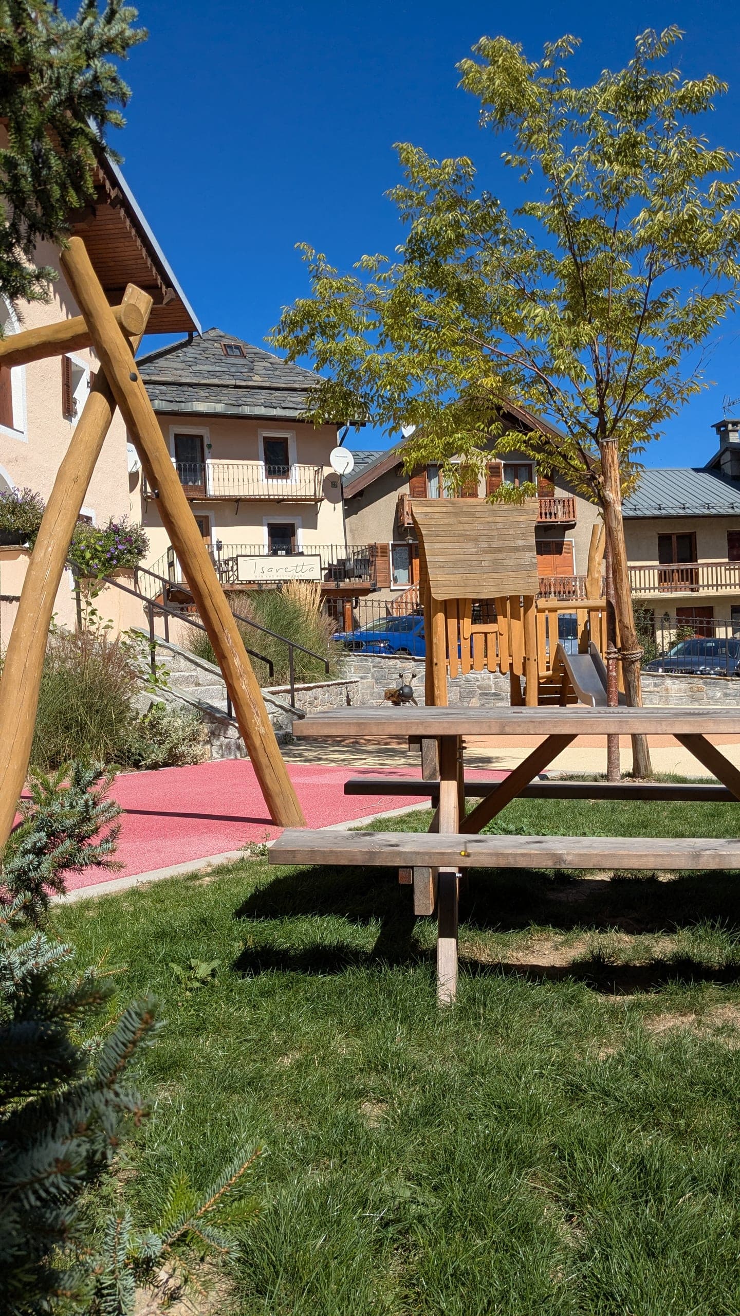 Le portique de balançoire en rondins au premier plan, une table de pique-nique en bois sur la pelouse, le sol souple rouge de l’aire de jeux derrière, les façades du village et un jeune arbre au feuillage clair.